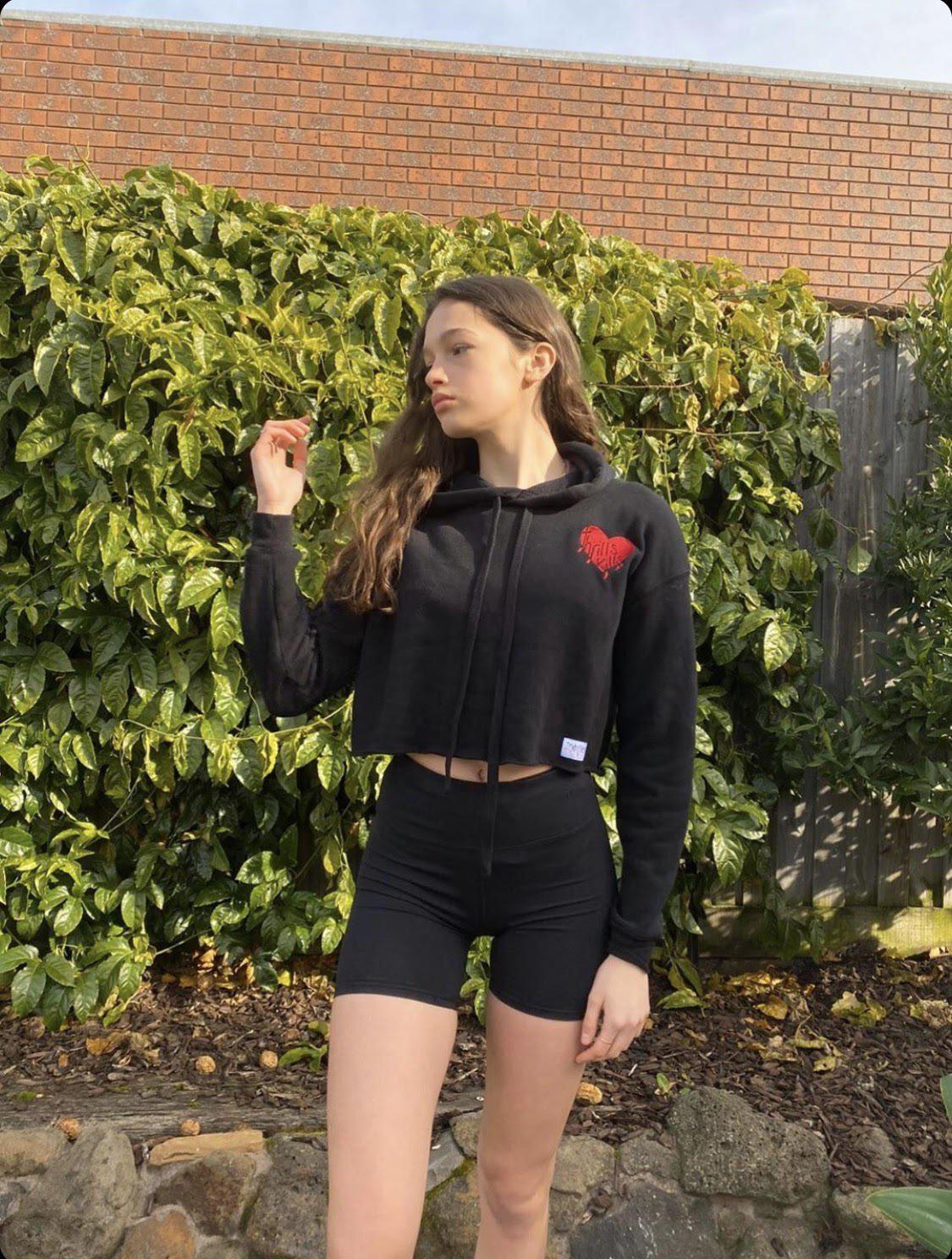 Model pictured showing off her long skinny legs is wearing a Supreme Black Embroidered Love Heart Crop Fleece Hoodie in a garden with green plants in background