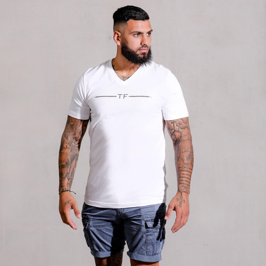 Muscle hunk with tattoos and beard modelling wearing White V Neck Signature Escape T-shirt and jeans for a causal look. Printed on the chest is the feels apparel signature slogan.
